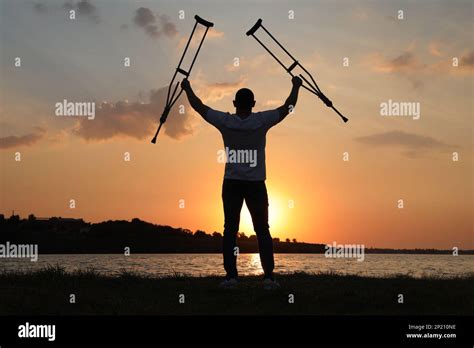 Man Raising Hands With Underarm Crutches Up To Sky Near River At Sunset
