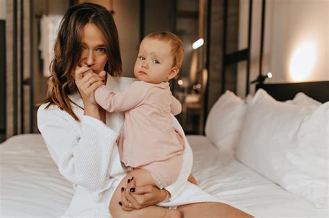 목욕 가운에 녹색 눈을 가진 여자는 그녀의 아이의 손에 키스 엄마와 딸 집에서 침대에 앉아 포즈 무료 사진