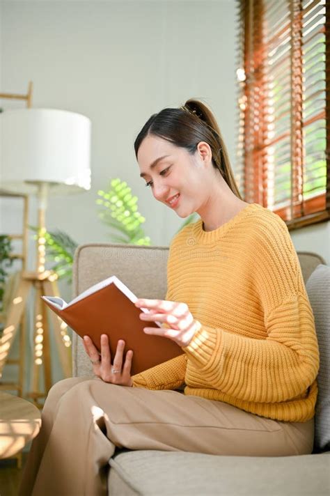 Attractive Asian Woman In Cozy Sweater Reading A Book On Sofa In Her