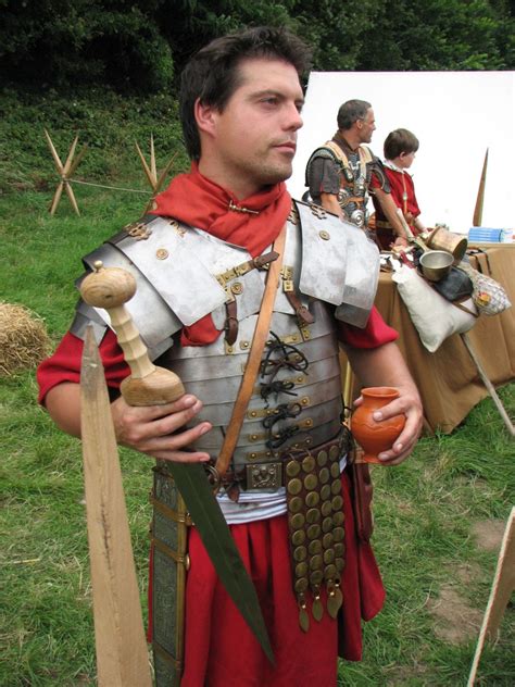 A Man Dressed In Roman Armor Holding Two Wooden Mallets And An Orange