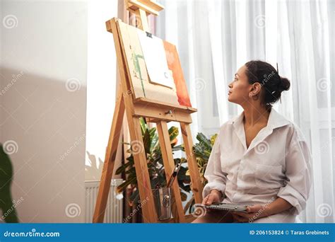 Woman Painter In White Shirt Standing In Front Of Easel Thinking And