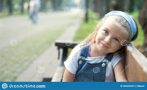 Small Happy Child Girl Sitting On A Bench Resting In Summer Park Stock