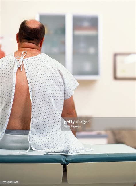 Mature Man In Medical Gown Sitting On Exam Table Rear View Photo