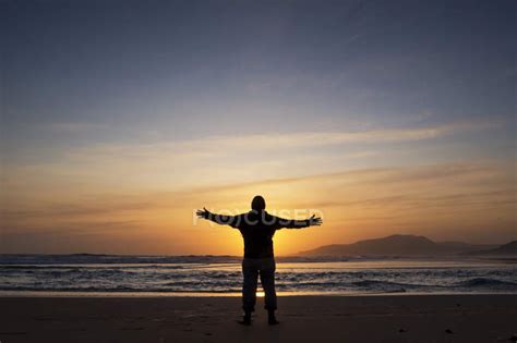 Silhouette Of Man Standing With Arms Outstretched On Beach At Sunset