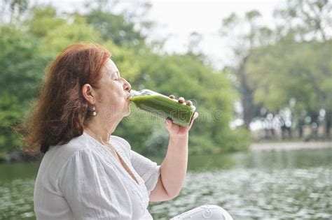 Mature Hispanic Woman Sitting In A Park Drinking Green Juice From A