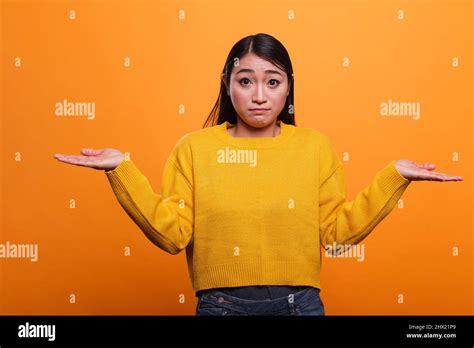 Confused Uncertain Asian Woman Shruging Unsurely While Wearing Yellow Sweater On Orange