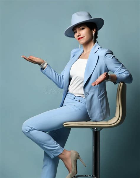 Smiling Short Haired Brunette Woman In Blue Business Suit And Hat Sits On Stool Holding Hand Up