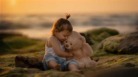 Cute Baby Is Sitting On Sand Hugging Toy Teddy Bear