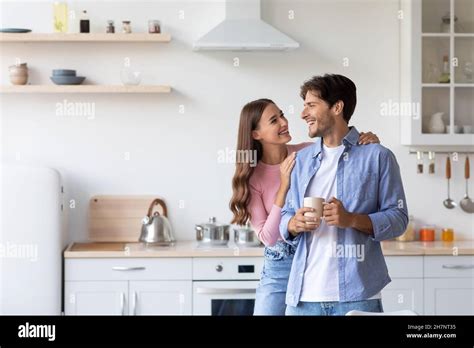 Cheerful Millennial European Wife Hugs Husband With Cup Of Hot Drink In Minimalist Kitchen