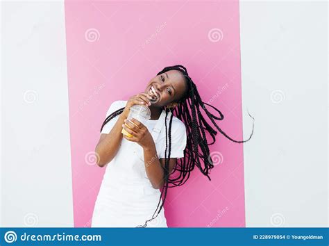 cheerful woman with braids drinking a juice and looking at camera while standing at pink wall
