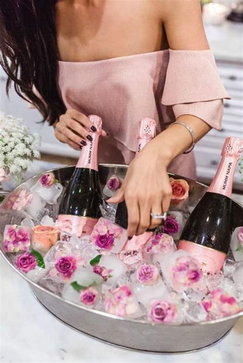A Woman Is Pouring Champagne Into An Ice Bucket With Pink Flowers On The Side And Two Bottles In It
