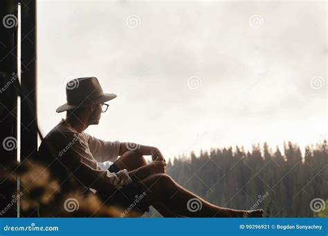 Hipster Man Sitting On Porch Of Wooden House Looking At Woods I Stock
