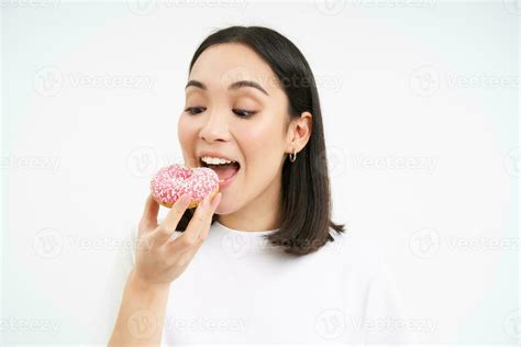 Close Up Of Smiling Pleased Asian Woman Eats Glazed Pink Doughnut Enjoys Eating Tasty Donnut
