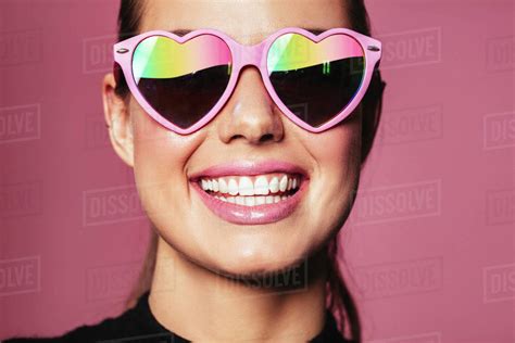 Closeup Portrait Of Beautiful Young Woman Wearing Heart Shaped Sunglasses And Smiling Against