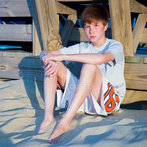 Young Boy Sitting On Sandy Beach And Leaning Against Boardwalk