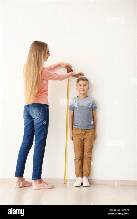 Young Woman Measuring Height Of Little Boy Near Light Wall Stock Photo