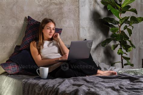 femme assise sur le lit avec un ordinateur portable sur ses genoux et une tasse blanche de côté