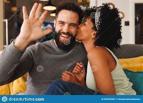 Happy Bearded Biracial Man Waving Hand And Girlfriend Kissing Him On Cheek While Relaxing On
