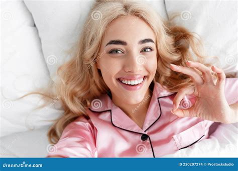 Cheerful Millennial European Blonde Female In Pink Pajama Shows Ok Gesture With Hand Lies On