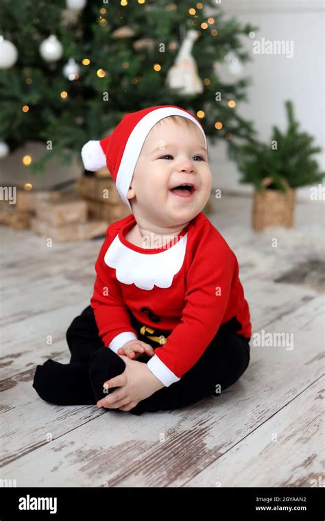 Baby Boy In Santa Claus Costume Is Sitting On The Floor And Laughing