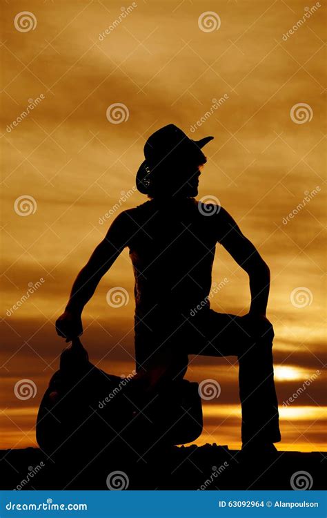 Silhouette Of Cowboy Kneeling By Saddle Look To Side Stock Photo