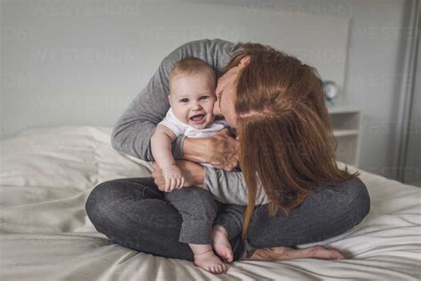 Happy Mother Kissing Cute Son While Sitting On Bed Against Wall At Home