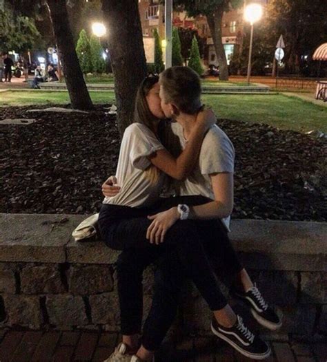 Two People Sitting On A Stone Bench Kissing Each Other In Front Of A Park At Night