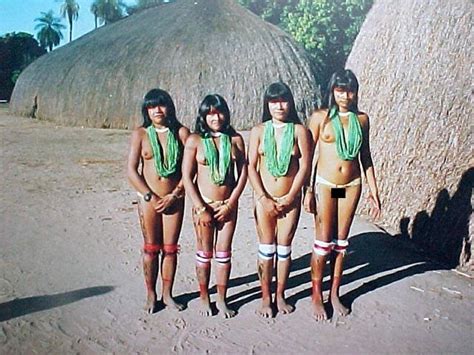 Four Women In Bathing Suits Standing Next To Each Other On The Beach With Grass Huts Behind Them