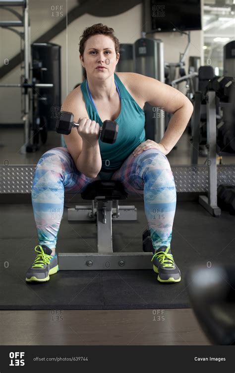 Full Length Of Woman Lifting Dumbbell While Sitting On Bench In Gym