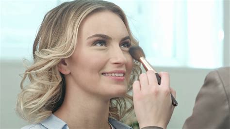 Applying Blush Makeup With Brush To Cheekbones Of Smiling Young Woman