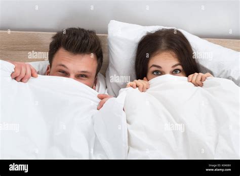Portrait Of Young Couple Hiding Face With Blanket In Bedroom Stock