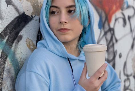 Blue Haired Teenage Girl In Light Blue Oversize Hoodie Staying Near Graffiti Wall With Coffee To