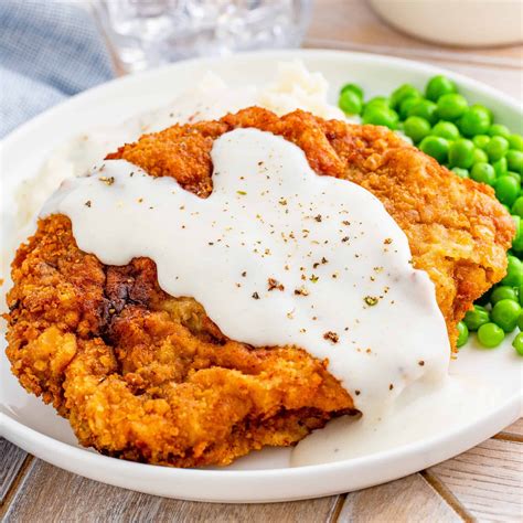 A White Plate Topped With Fried Chicken Covered In Gravy Next To Green Peas