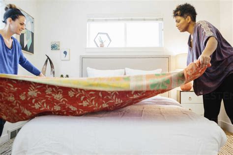 Two Women Making Up A Bed Spreading A Patterned Quilt Over The Double Bed In A Light Airy