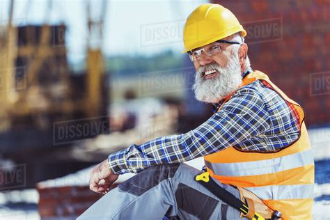 Bearded Worker In Reflective Vest And Hardhat Sitting At Construction