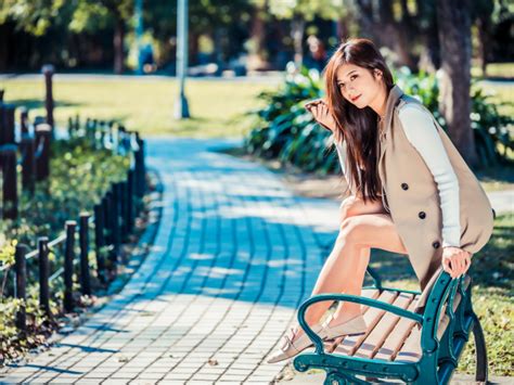 Молодая девушка азиатка сидит в пальто на лавке в парке обои для