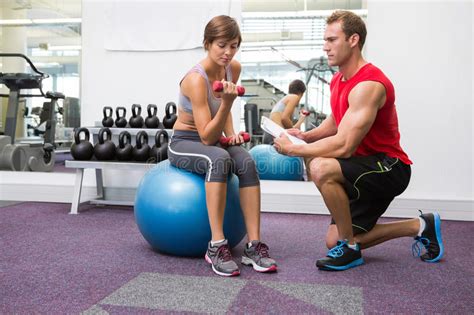 Personal Trainer With Client Sitting On Exercise Ball Lifting Dumbbell
