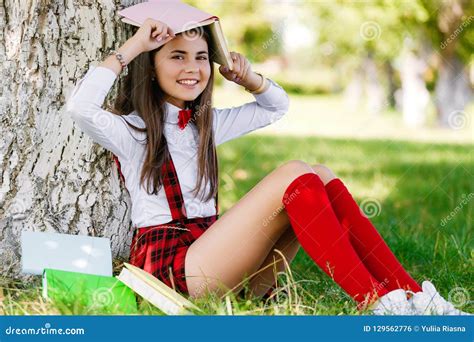 Young Schoolgirl In School Uniform Sitting On The Grass Under A Tree