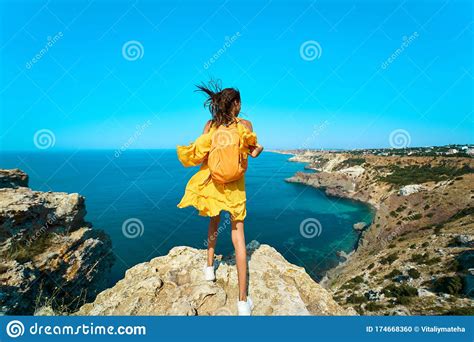 Rear View Of Traveler Woman Standing On Top Rock Beach With Backpack Bright Yellow Dress And