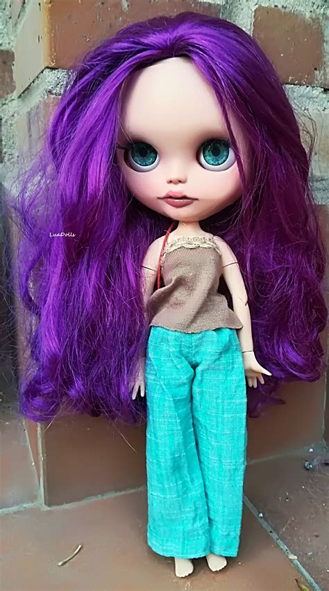 A Close Up Of A Doll With Purple Hair And Blue Pants On The Ground Next To A Brick Wall