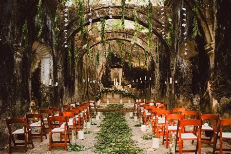 An Outdoor Ceremony Setup With Orange Chairs And Greenery On The Aisle