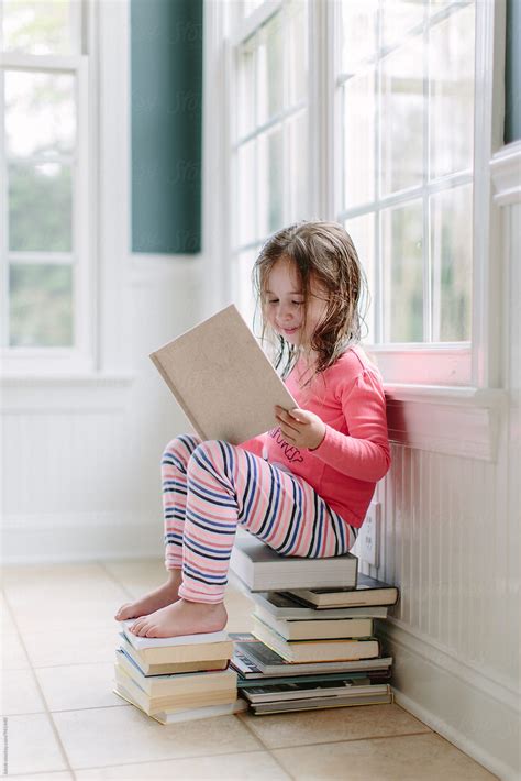 Cute Young Girl Sitting On A Stack Of Books Reading By Stocksy