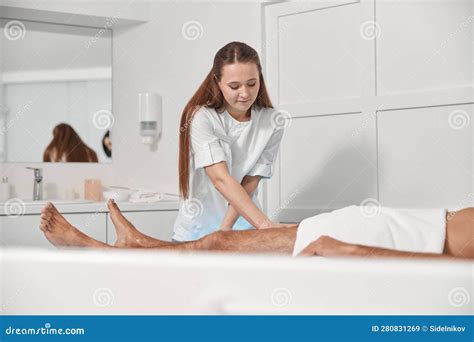 Smiling Woman Massages Legs Of Mature Man Lying On Couch In Clinic