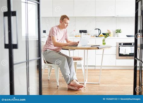 Young Casual Man Sitting By Table In The Kitchen In Front Of Laptop