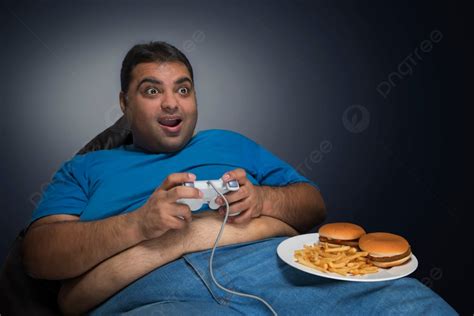 رجل سمين بطن مرئية من قميصه جالسًا يلعب لعبة فيديو مع طبق من البرغر والبطاطا المقلية على فخذه