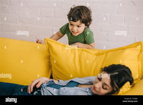Little Boy Standing Near Tired Mother Lying On Yellow Sofa In Living
