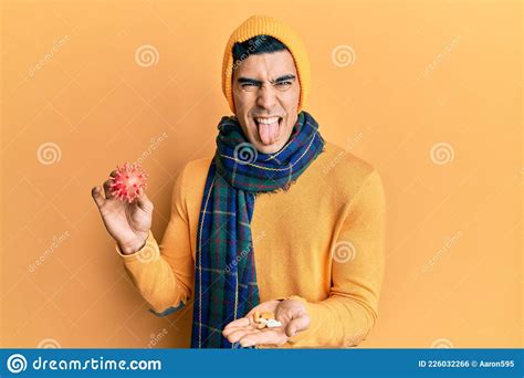 Handsome Hispanic Man Holding Virus Toy And Pills Sticking Tongue Out Happy With Funny