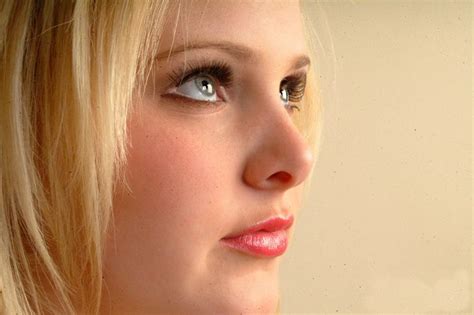 A Woman With Blonde Hair And Blue Eyes Looks Off Into The Distance While Wearing A Necklace
