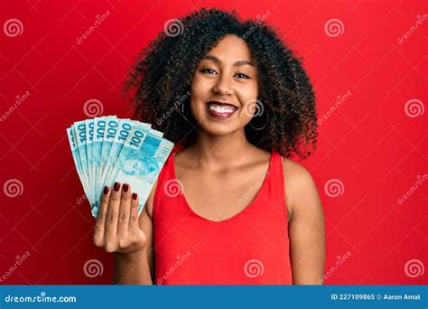 beautiful african american woman with afro hair holding 100 brazilian real banknotes looking