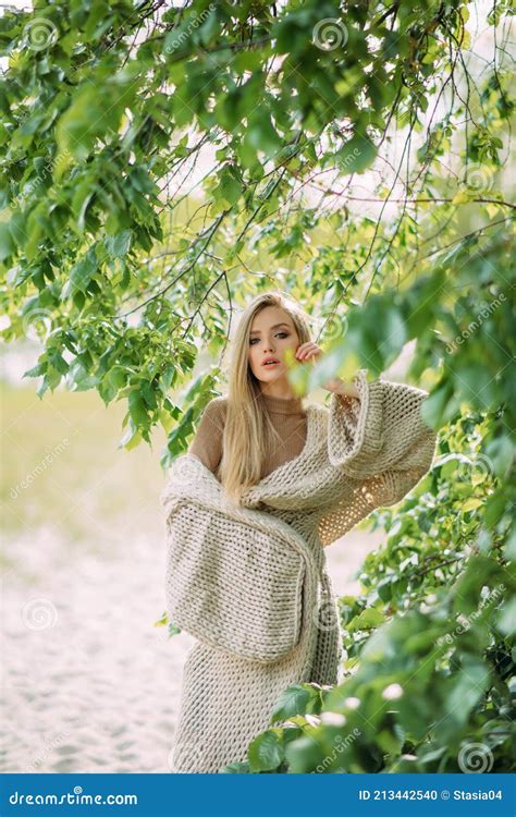 Blonde Woman Poses Among Green Branches Of Tree Near Lake Stock Photo